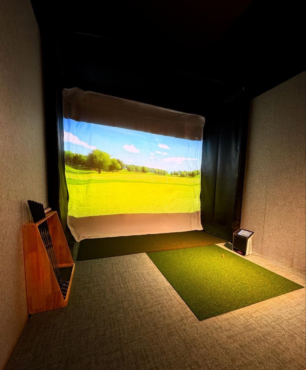 A wooden rack of irons standing beside the hitting mat in a simulator bay, with the projection screen lit up behind it