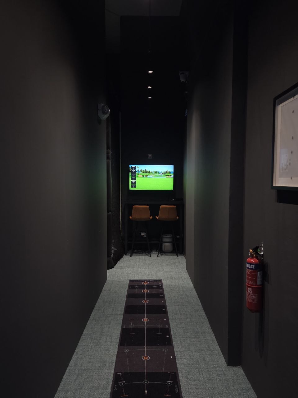 The putting lane — a practice putting mat marked with target rings running the length of a dark corridor toward a wall screen and bar seating