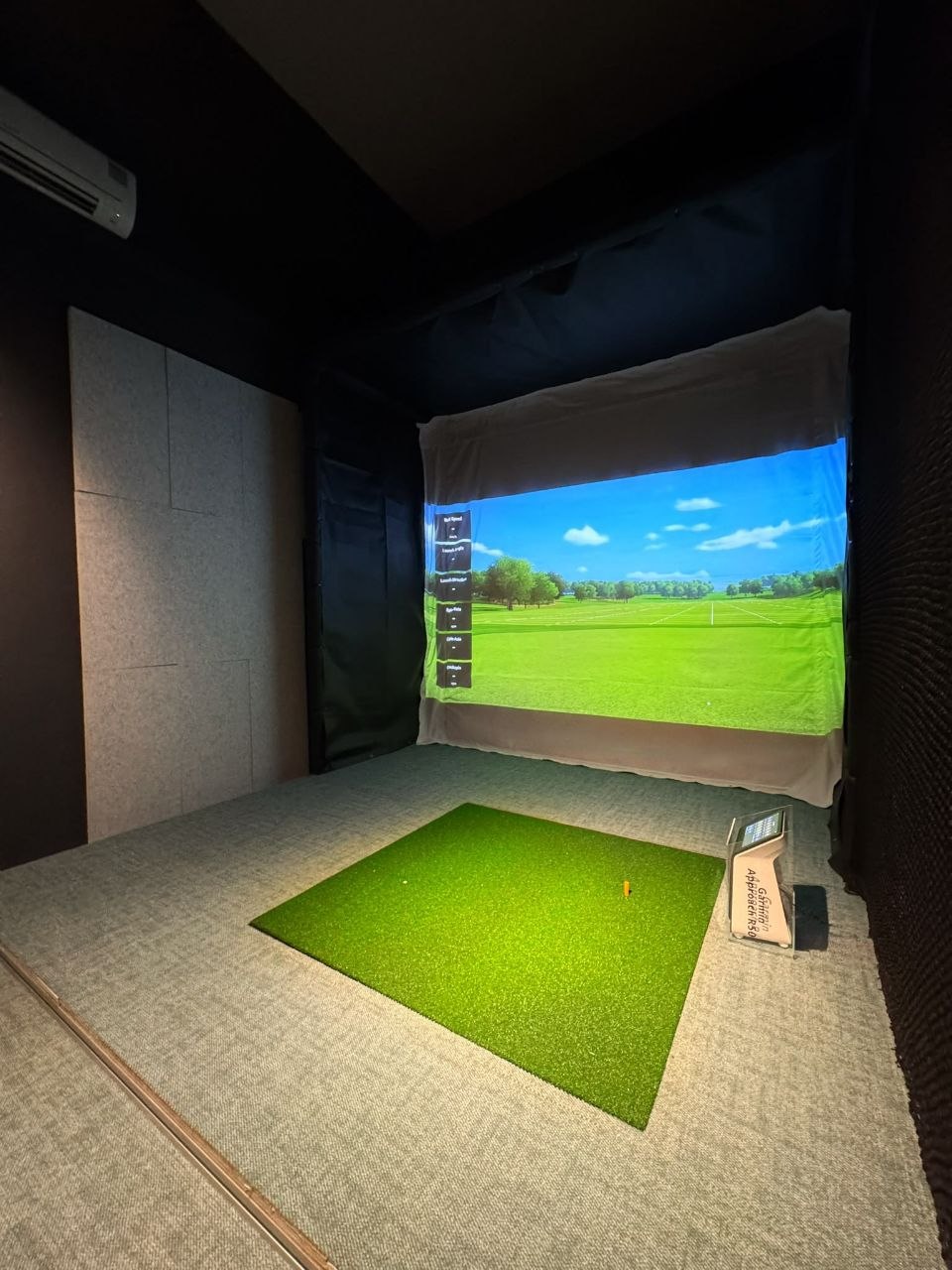 A simulator bay ready to play — a green hitting mat facing a full-width projection screen showing an open fairway, with a launch monitor set up beside the mat