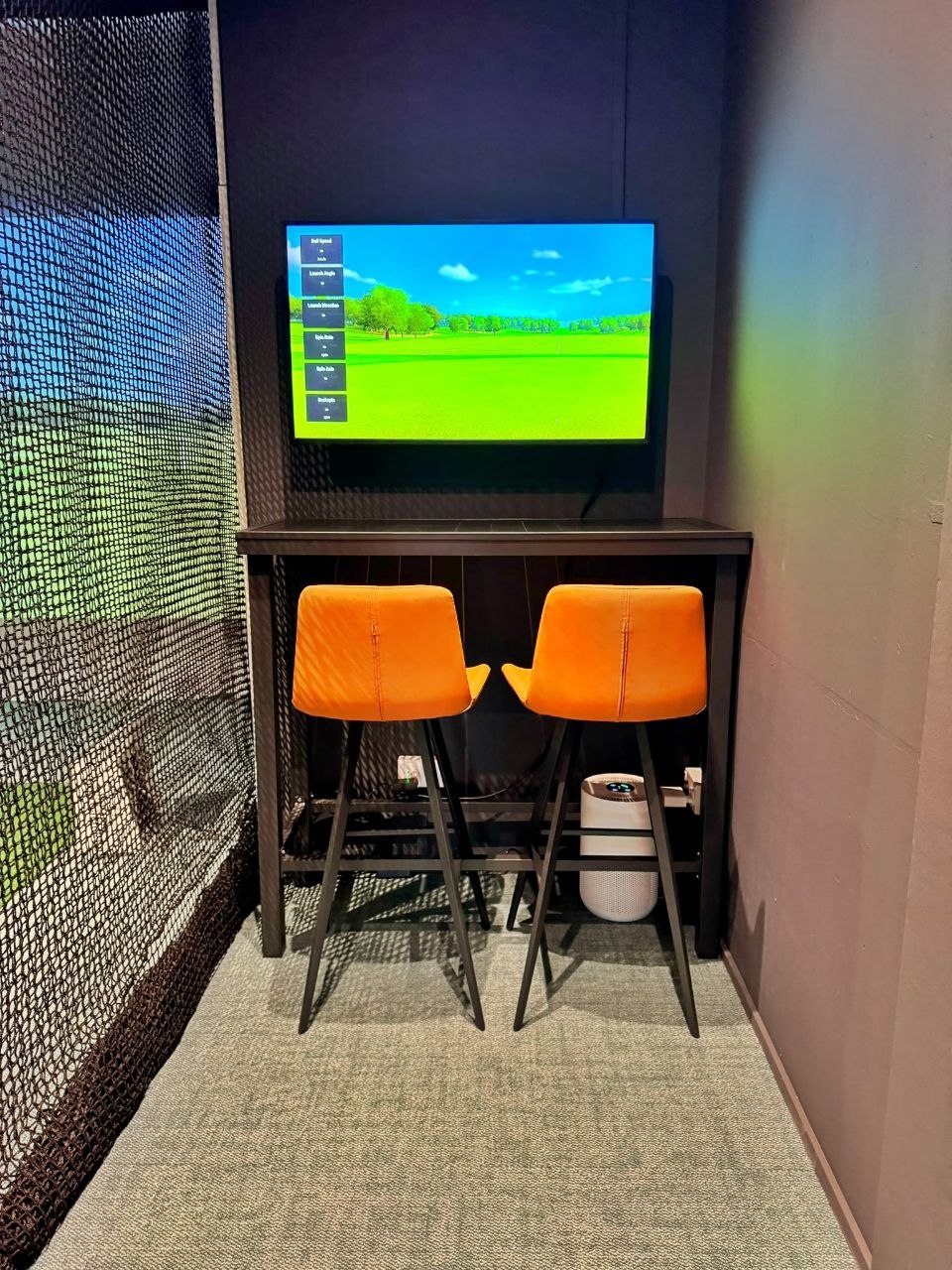 Two orange bar stools at a high table beneath a wall-mounted screen, next to the netting of a hitting bay
